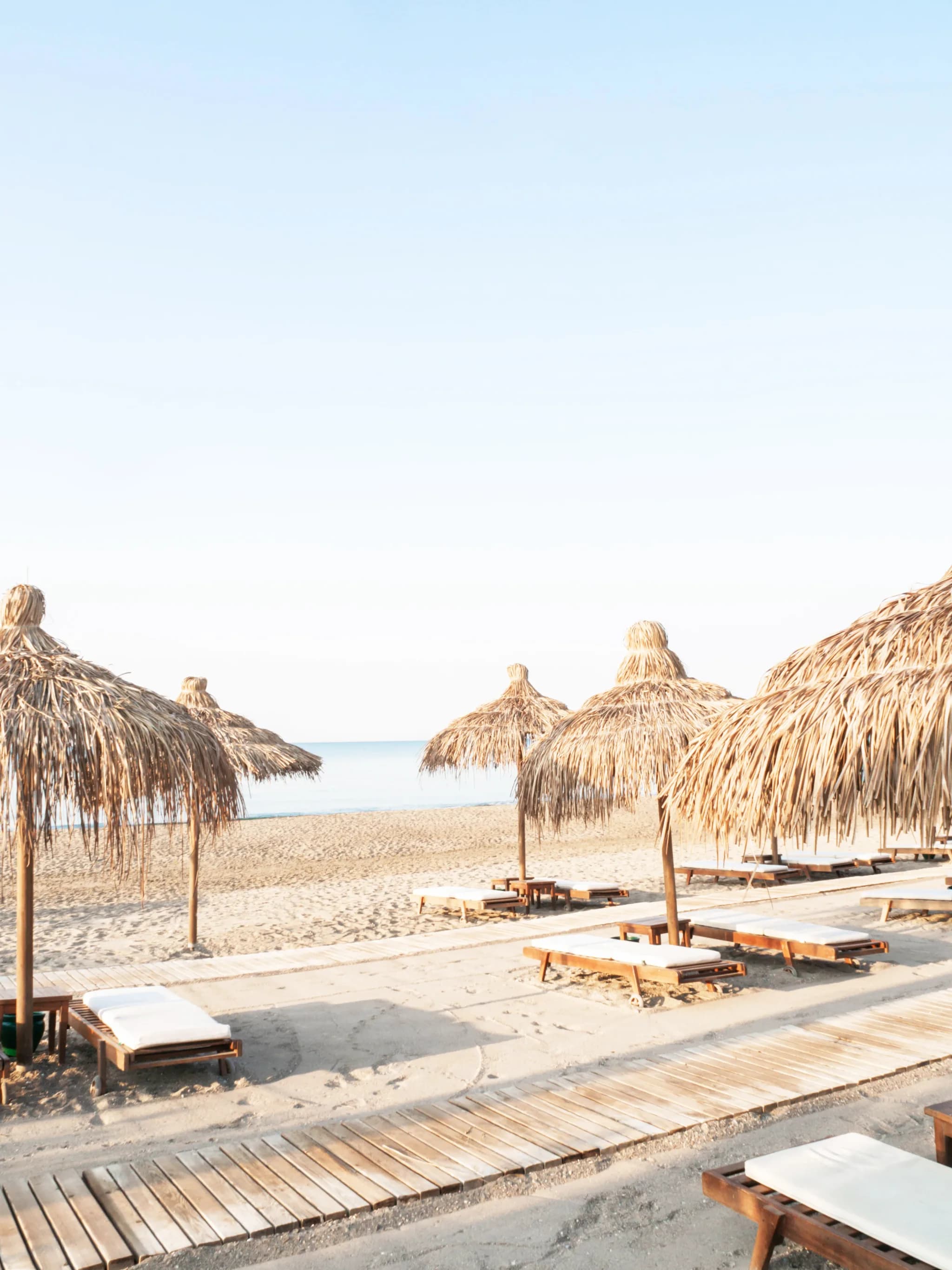 Empty beach loungers under palm-thatched umbrellas at sunrise, awaiting guests