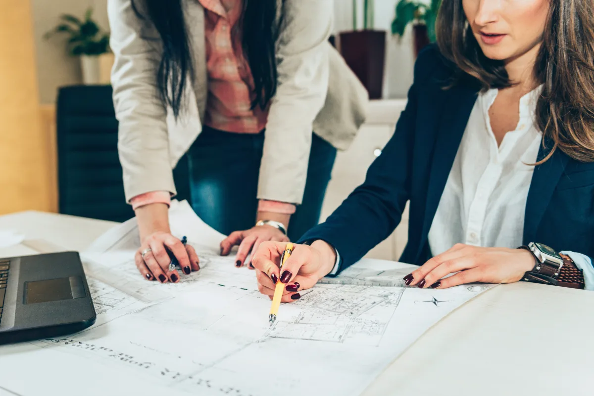 Two women in blazers reviewing architectural plans at a shared desk with a laptop